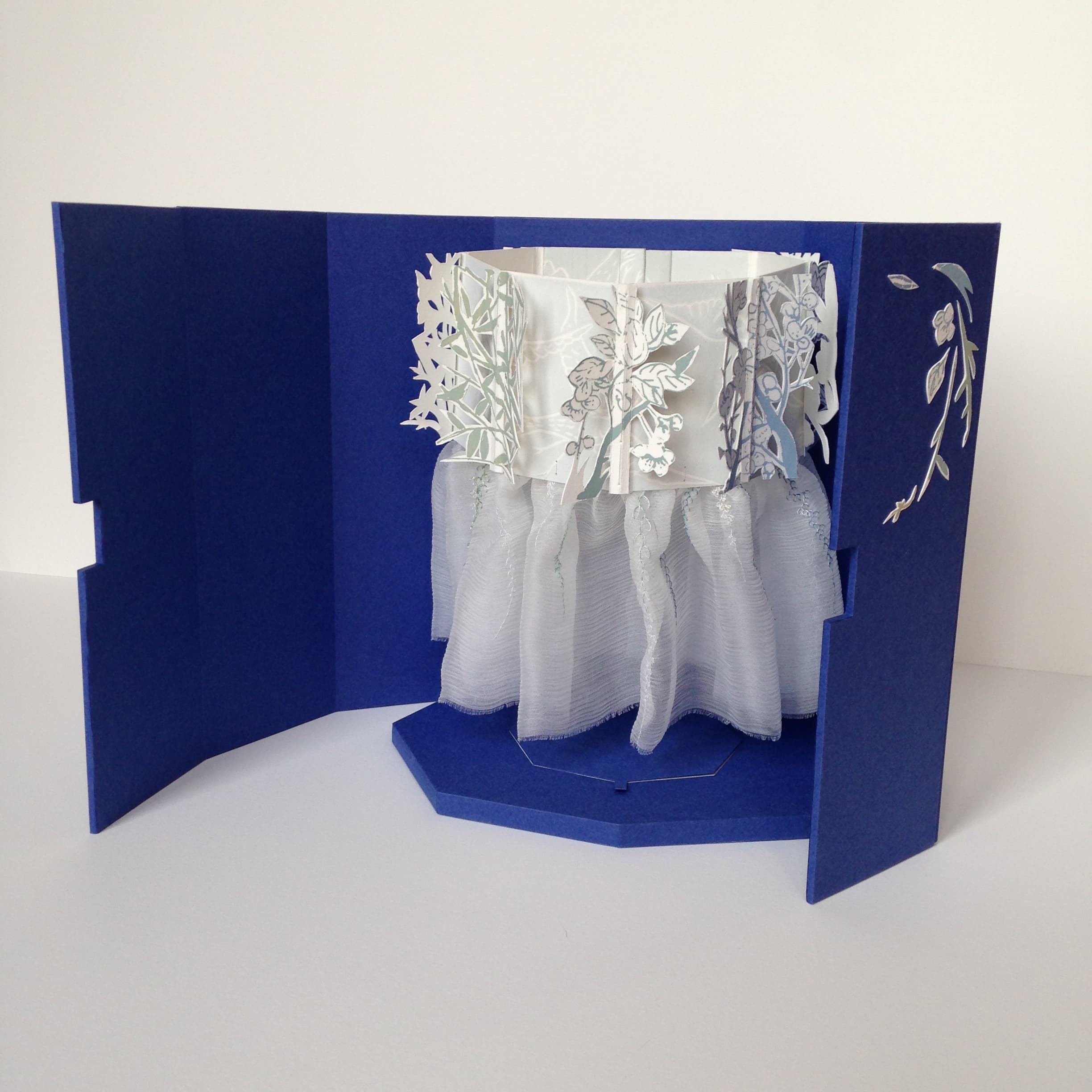 Boîte facettée ouverte. Livre objet fait de découpes papier, de sérigraphie et de textile brodé, suspendu à la boîte.