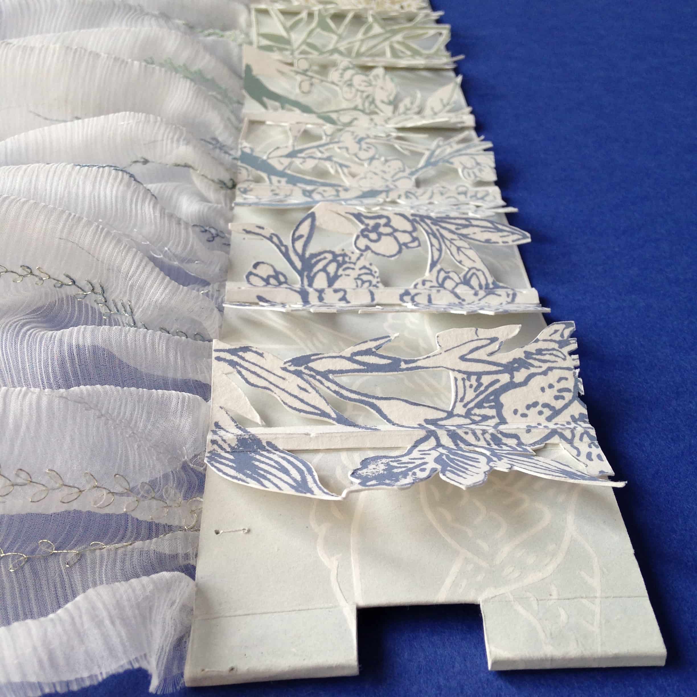 Livre d'artiste mis à plat. Vue sur les cahiers sérigraphiés et leurs découpes papier ainsi que sur une bande de textile en soie brodée au point d'épine.