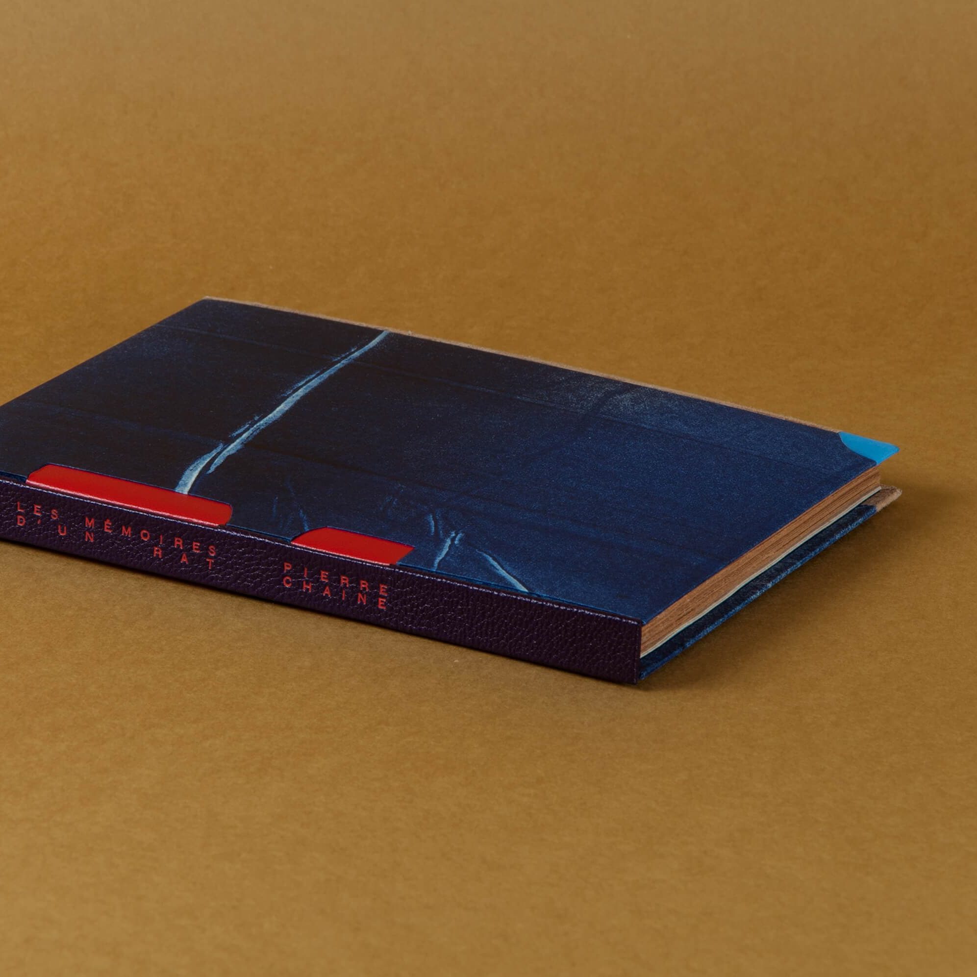 Reliure posée à plat laissant voir le dos en cuir violet, les rubans rouge, le plat avant en papier imprimé et une tranche